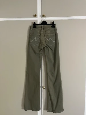 Bootcut byxor från Nelly i khaki - Bootcut byxor från Nelly i khakigrön färg med vita kontrastsömmar och dubbla knappar fram. Snygga fickor bak med dekorativa detaljer och klassisk femficksdesign. Materialet är bomull och byxorna har en lågmidjad passform.