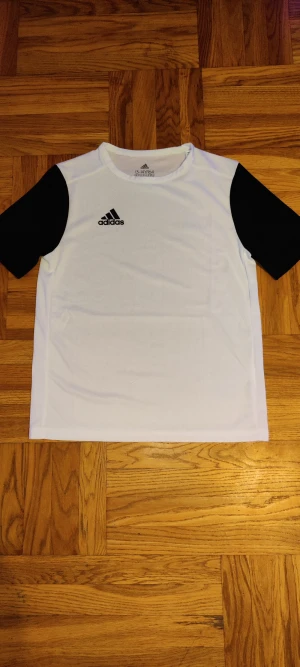 Vit och svart Adidas t-shirt - Snygg t-shirt från Adidas i vitt med svarta ärmar och klassisk logga på bröstet. Tillverkad i ett lätt och sportigt polyestermaterial som andas. Perfekt för dig som gillar en stilren och sportig look.
