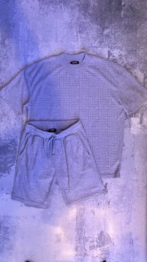 Grå sett shorts och t shirt med mönster från BohooMAN - Säljer ett snyggt sett grå shorts och t shirt från bohooMAN med snyggt geometriskt mönster i tyget. Shortsen har snörning i midjan och är gjorda i ett mjukt material som känns riktigt skönt mot huden. Perfekta för chill dagar eller när du vill ha en avslappnad stil.