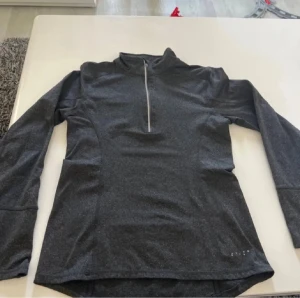 Mörkgrå långärmad träningströja - Snygg mörkgrå långärmad träningströja från Craft med half zip framtill. Perfekt för löpning eller gym, med skön passform och lätt material som andas. Tröjan har en hög krage och diskret logga nere vid höger sida.