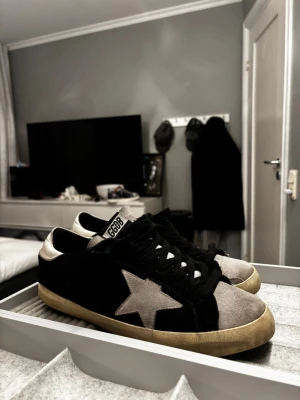Golden Goose - Snygga Golden goose sneakers i svart och grå mocka med en ikonisk stjärndetalj på sidan. Skorna har snörning, rund tå och en beige platt sula. Silverfärgad häl och GGDB-logga på tungan. Kommer med originalkartong och dustbag.