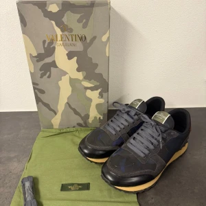 Valentino Garavani Rockrunner sneakers - Snygga Valentino Garavani Rockrunner sneakers med camouflagemönster i mörkblått och grått, röda detaljer på hälen och beige mellansula. Skorna har snörning, platt sula med nitar och är tillverkade i mocka, skinn och textil. Perfekt för dig som gillar exklusiva och unika sneakers. Storlek 43