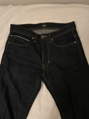 Mörkblå jeans från Neuw - Original pris 1400 kr. Snygga mörkblå jeans från Neuw med klassisk femficksdesign och kontrasterande gul söm. Jeansen har rak passform, normal midja och detaljer som metallring och mönster på bakfickan och fram. Materialet är robust denim i bomull. Midjemått:38 cm Innerbenslängd:78 cm 
