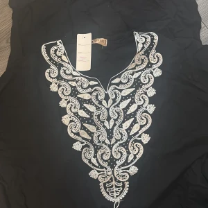 Svart långklänning med broderi från Styley - Svart långklänning med v-ringning och detaljerat silvrigt broderi framtill. Klänningen har en dekorativ tofs och ett unikt mönster längs halsringningen. Perfekt för dig som vill sticka ut med eleganta detaljer och snygg passform.