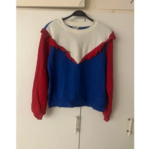 Färgglad tröja med volang från Monki - Säljer en unik tröja från Monki i storlek L med cool färgblockning i blått, rött och vitt. Tröjan har långärmade röda ärmar, blå kropp och vit överdel med röd volangdetalj framtill. Perfekt för dig som gillar statement-plagg nu till jul och vill sticka ut. En jultröja som kan användas året runt. 