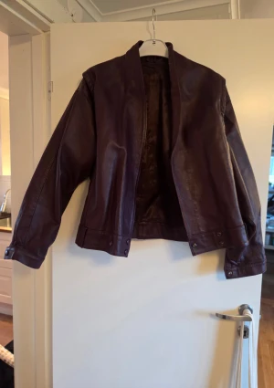 Lila/brun skinnjacka, storlek L - Snygg skinnjacka i lila ton med loose passform och stilrena detaljer. Jackan har en öppen front, bred krage och knappar vid nederkanten. Insidan är gjord med ett glansigt material som känns lyxigt. Storlekslappen har fallit bort, men det är en storlek L. Endast använd vid ett tillfälle. 