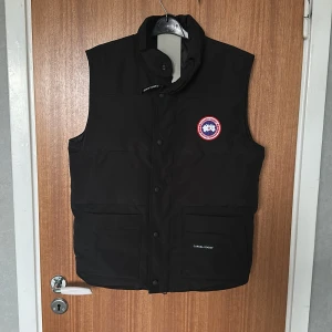 Svart dunväst från Canada Goose - Säljer en svart dunväst från Canada Goose med klassisk logga på bröstet och hög krage. Västen har knappar framtill och två stora fickor. Perfekt för lager-på-lager och stilren look.
