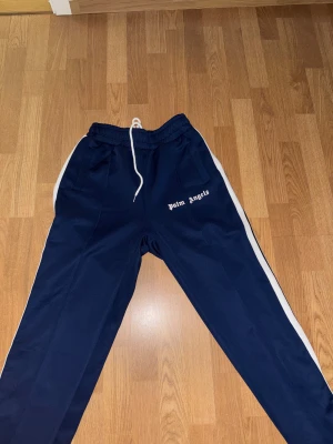 Palm Angels blå mjukisbyxor med vita ränder - Snygga blå mjukisbyxor från Palm Angels med vita sidoremsor och logga broderad på framsidan. Byxorna har elastisk midja med vit snörning och raka ben. Materialet är mjukt och har en lätt glansig finish, perfekt för en chill och sportig look.