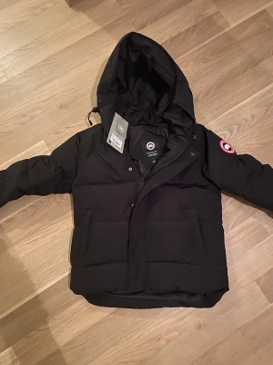 Svart dunjacka från Canada Goose - Säljer en svart dunjacka från Canada Goose med huva och klassisk logga på ärmen. Jackan har stora fickor framtill, dragkedja och knappar. Perfekt för kalla dagar och riktigt snygg streetstil.