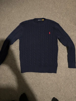 Mörkblå stickad tröja från Polo Ralph Lauren - Säljer en mörkblå stickad tröja från Polo Ralph Lauren med klassiskt kabelstickat mönster och rund hals. Tröjan har långärmad passform och en röd broderad logga på bröstet. Perfekt för dig som gillar stilrena och tidlösa plagg. Helt ny oanvänd.