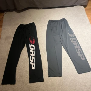 GASP gymbyxor svart och grå i size M - Två par mjukisbyxor från GASP, ena i svart med stor vit och röd logga längs benet, andra i mörkgrått med grå logga. Båda har elastisk midja och raka ben, tillverkade i mjukt material som passar perfekt för gym eller chill. Snyggt tryck längs benet ger en sportig vibe.
