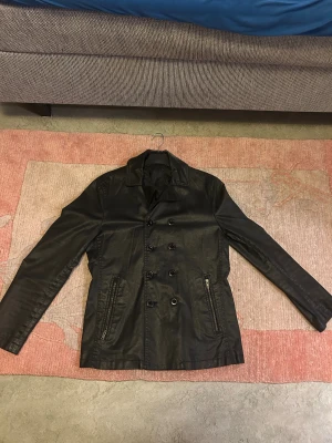 Tiger of sweden jacka - Tiger of Sweden – exklusiv herrjacka Jacka i skinnliknande Limonta-tyg (made in Italy). Klassisk dubbelknäppt modell med hög kvalitet och snygg passform. Lätt, slitstark och perfekt för vår/höst. Nypris uppskattningsvis ca 3 000–5 000 kr. • Storlek: L • Färg: Svart • Material: Limonta technical fabric