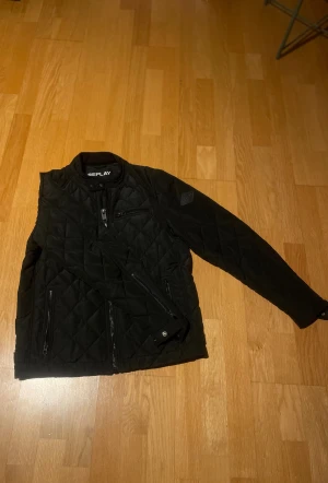 Svart quiltad jacka från Replay - Snygg svart quiltad jacka från Replay med dragkedjor på ärmar och framficka. Jackan har en stilren look med diskret logga på bröstet och flera praktiska fickor. Perfekt för dig som gillar en clean och modern stil. Storlek S