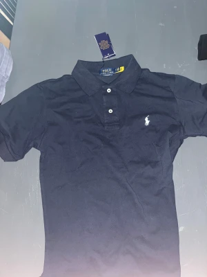 Svart pikétröja från Polo Ralph Lauren - Svart pikétröja från Polo Ralph Lauren i custom slim fit. Tröjan har klassisk krage, två vita knappar och den ikoniska broderade loggan på bröstet. Materialet är mjuk bomull som känns skönt mot huden. Perfekt för dig som gillar stilrena och tidlösa plagg.