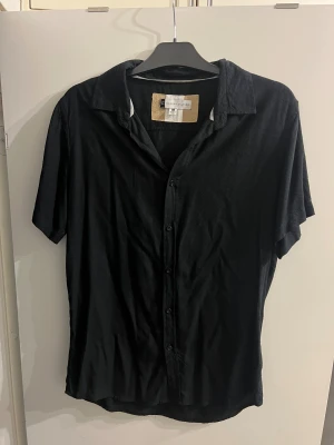 Svart skjorta från Modern Culture - Svart skjorta från Modern Culture i storlek M, tillverkad i 100% viskos. Skjortan har klassisk krage, korta ärmar och knappar framtill. Enkel och stilren design som funkar till många olika looks. Perfekt för dig som gillar minimalistisk stil.