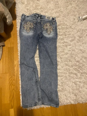 Blå bootcut jeans från miss me - Snygga blå bootcut jeans från miss me med coola broderade kors och paljettdetaljer på bakfickorna. Jeansen har normal passform, klassisk femficksdesign och silverfärgade knappar. Materialet är jeans med slitningar och ljusare partier för en trendig look.