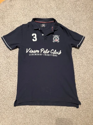 Svart pikétröja Vinson Polo Club S - Snygg Svart pikétröja från Vinson Polo Club med vit broderad logga och text framtill. Klassisk krage med knappar och vita detaljer på ärmsluten. Perfekt för en sportig och stilren look. 