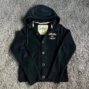 Mörk blå hoodie från Hollister med knappar - Svart hoodie från Hollister med broderad logga och text på bröstet. Tröjan har knäppning med stora knappar, dragsko i huvan och två fickor framtill. Mjuk insida och klassisk passform, perfekt för chill dagar.