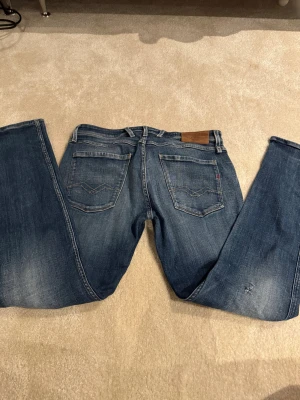 Replay Anbass jeans blå slim fit.      2.  W-33 L-32 - Snygga blå Replay Anbass jeans i slim fit med klassisk femficksdesign och dragkedja. Jeansen har en lätt tvättad look med subtila slitningar och kontrastsömmar. Passar dig som gillar en modern och stilren denimstil.