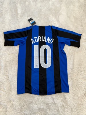Inter Adriano #10 Nike fotbollströja - Säljer en klassisk Inter Milan fotbollströja med Adriano och nummer 10 på ryggen. Tröjan är blå och svart med breda vertikala ränder, Pirelli-logga på bröstet och Nike Swoosh. Materialet är lätt och andas, perfekt för match eller träning.