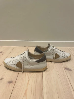 Philippe Model Skor - Snygga vita sneakers från Philippe Model med beige mockadetalj på sidan och bak, samt mörkgrå häl. Klassisk låg modell med rund tå, platt sula och snörning. Skorna är tillverkade i skinn och mocka, vilket ger en lyxig känsla och cool kontrast.           Storleken på skorna är 39. 