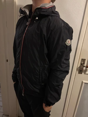 Marinblå vindjacka från Moncler med huva - Marinblå vindjacka från Moncler med huva och dragsko, snygga röd-vita detaljer längs dragkedjan och logga på ärmen. Jackan har hel dragkedja framtill och grått innerfoder vid kragen. Perfekt för dig som vill ha en stilren och sportig look.