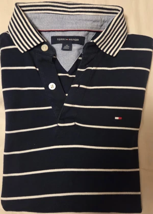 Marinblå randig pikétröja Tommy Hilfiger - Snygg marinblå pikétröja från Tommy Hilfiger med vita ränder och klassisk krage med kontrasterande ränder. Tröjan har korta ärmar, två knappar vid halsen och diskret logga på bröstet. Perfekt för dig som gillar preppy stil.