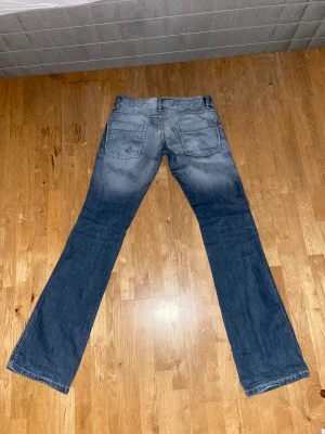 Lågmidjade bootcut jeans från Diesel - Säljer ett par blå bootcut jeans från Diesel med snyggt slitna detaljer och klassisk femficksdesign. Jeansen har en ljus tvätt med kontrasterande partier och en extra ficka med lock på ena benet. Perfekt för dig som gillar en avslappnad och cool stil.