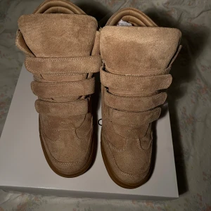 Beige wedge sneakers med kardborre - Snygga beige sneakers med inbyggd kilklack och flera breda kardborreband över foten. Skorna har en rund tå, platt gummisula och är tillverkade i mocka för en mjuk och lyxig känsla. Perfekt för dig som vill ha lite extra höjd men ändå behålla en sportig vibe.
