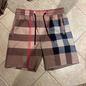 Burberry shorts  - Snygga shorts från Burberry med klassiskt rutigt mönster i beige, brunt, svart, vitt och rött. De har elastisk midja med svart snörning och praktiska fickor. Perfekta för sommaren och ger en lyxig vibe till din outfit.