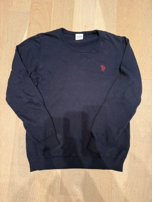 Marinblå stickad tröja från US Polo Assn - Snygg marinblå stickad tröja från US Polo Assn med röd broderad logga på bröstet. Rund halsringning och ribbade muddar vid ärmslut och nederkant. Perfekt för dig som gillar klassisk stil med en sportig touch.