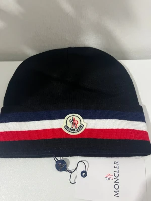 Moncler mössa med ränder - Snygg svart Moncler mössa med breda randiga detaljer i blått, vitt och rött. Märkets logga framtill ger en exklusiv touch. Mössan är stickad och har en klassisk form som passar perfekt för kalla dagar. Helt ny 