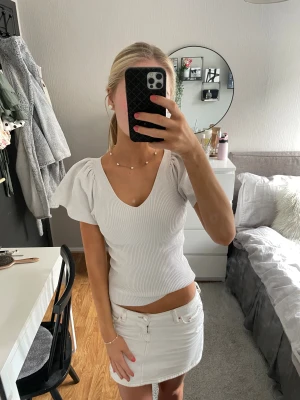 Vit ribbad topp med puffärm - Supersöt vit ribbad topp med puffiga korta ärmar och v-ringning. Toppen har öppen rygg med snygg knyt-detalj och sitter figurnära. Perfekt till jeans eller kjol för en fräsch look. Materialet känns mjukt och stretchigt.