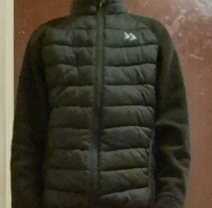 Svart quiltad pufferjacka från Everest - Svart pufferjacka från Everest med quiltad framsida och rygg, samt släta ärmar. Jackan har dragkedja hela vägen och en liten logga på bröstet. Perfekt för kyliga dagar och har en clean, sportig look som funkar till streetwear.