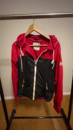 Svart vindjacka från Moncler - Snygg vindjacka från Moncler i rött och svart med vit dragkedja och snörning i huvan. Jackan har två fickor med dragkedja och Moncler-logga på ärmen. Perfekt för dig som gillar streetstyle och vill ha något som sticker ut.