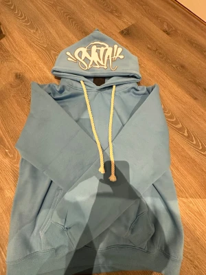 Ljusblå hoodie från syna world  - Säljer en ljusblå hoodie från syna world med coolt graffiti-tryck på huvan och vita snören. Tröjan har känguruficka framtill och mjukt material som känns skönt mot huden. Perfekt för dig som gillar streetwear och vill sticka ut med en unik look.