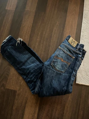 Mörkblå jeans från Nudie Jeans med slitningar - Säljer ett par mörkblå jeans från Nudie Jeans med klassisk femficksdesign och orangea sömmar samt slitningar där nere. Modellen ”Grim Tim”, W30 L34.