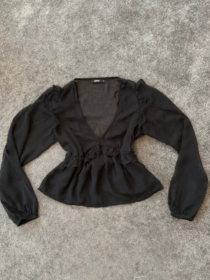 Svart blus med volang - Svart blus från Bik Bok med djup v-ringning och långa ärmar. Blusen har volangdetaljer vid axlar och midja som ger en cool och feminin touch. Perfekt för dig som gillar stilrena och trendiga plagg.
