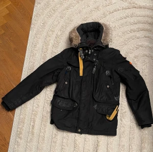 Parajumpers svart parkajacka med päls - Svart parkajacka från Parajumpers med äkta päls på huvan, cool gul rem och flera fickor med dragkedja och knappar. Perfekt för kalla dagar. Det går även att ta av fodret på insidan för att kunna använda jackan på höst/vår. Jackan är i bra skick och väl omhändertagen. Kvitto finns. 