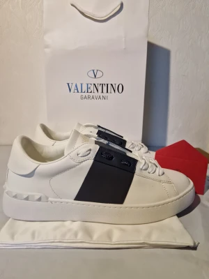 Valentino Garavani vita sneakers - Snygga vita Valentino Garavani sneakers med bred svart panel på sidan och coola nitar längs sulan. Skorna har rund tå, platt sula och vita snören. Tillverkade i skinn för en lyxig känsla och stilren look. Perfekta för dig som gillar exklusiva märken och modern design.
