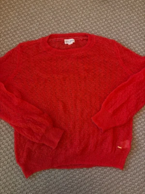 Röd stickad tröja Holly & Whyte - Säljer en röd stickad tröja från Holly & Whyte by Lindex i storlek M. Tröjan har ett luftigt, spetsliknande mönster och ribbade muddar vid ärmslut och nederkant. Perfekt för lager-på-lager och ger en färgklick till din outfit.