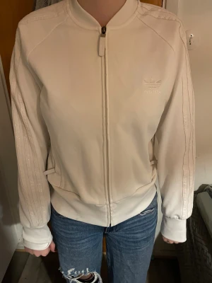 Vit bomberjacka från Adidas - Säljer en fräsch vit bomberjacka från Adidas med klassiska ton-i-ton ränder på ärmarna och broderad logga på bröstet. Jackan har dragkedja framtill, ribbade muddar och en clean, sportig look som passar perfekt till jeans eller joggers.