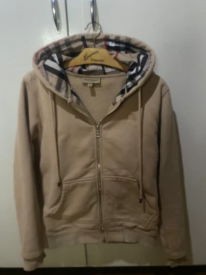 Beige hoodie från Burberry med dragkedja - Säljer en beige hoodie från Burberry med klassiskt rutigt foder i huvan. Tröjan har dragkedja framtill, snörning i huvan och två fickor. Perfekt för dig som vill ha en stilren och lyxig vardagslook med ikoniska Burberry-detaljer.