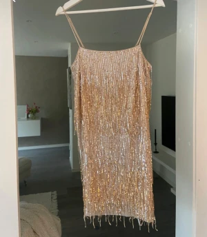 Champagnefärgad paljettklänning från H&M - Champagnefärgad kortklänning från H&M med tunna axelband och glittriga paljetter över hela klänningen. Klänningen har en rak passform och är perfekt för dig som vill sticka ut med en glittrig look. Materialet är polyester och ger en lyxig känsla.