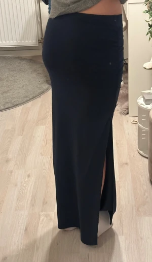 Mörkblå långkjol med slits och rynk - Säljer en svart långkjol med tight passform och medelhög midja. Kjolen har en snygg slits på sidan och rynkade detaljer upptill som ger en cool och trendig look. Materialet är stretchigt och följsamt, perfekt för att skapa en stilren siluett.