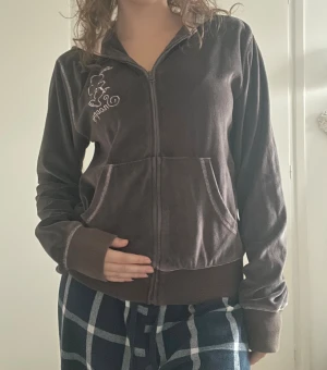 Brun Snoopy kofta i sammet - Mjuk brun kofta med dragkedja och luva, gjord i sammet. På bröstet finns ett broderat Snoopy-motiv i vitt. Den har långa ärmar, ribbade muddar och två fickor framtill. Den är även rosa på insidan av vissa delar (exempelvis luvan). Väldigt bekväm!🙌