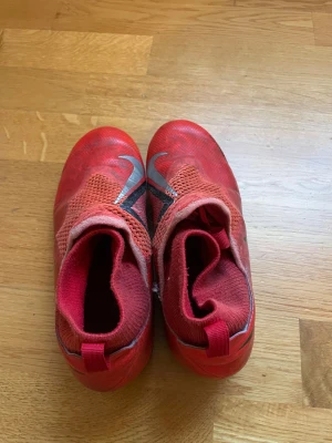Röda Nike fotbollsskor strl 35 - Säljer ett par röda Nike fotbollsskor med svart swoosh och mönstrad ovandel i syntetmaterial. Skorna har rund tå, platt sula med dobbar och meshpartier för extra ventilation. Perfekt för dig som vill ha snygga och funktionella skor på planen.