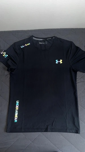 Svart Under Armour t-shirt för träning - Svart t-shirt från Under Armour med färgglada och reflekterande loggor och textdetaljer på bröst, axel och nederkant. Står storlek M men passar S, kom dm för fler bilder eller frågor !