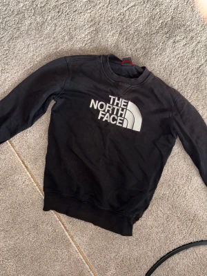 Svart sweatshirt från The North Face - Svart sweatshirt från The North Face med stort vitt logotryck på bröstet och mindre logga på ryggen vid axeln. Tröjan har rund hals, långa ärmar och är gjord i mjuk bomull. Perfekt för chill dagar eller när du vill ha en clean streetstyle.