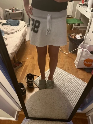 Grå sportig kjol med snörning - Grå kortkjol i sweatshirtmaterial med bred resår i midjan och vit snörning framtill. Tryckt svart siffra '95' på ena sidan ger en sportig vibe. Kjolen har inbyggda shorts under för extra komfort och rörelsefrihet.